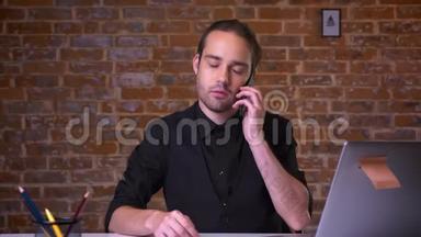 漂亮的白种人商人坐在办公桌前冷着身子，挂在电话上看着手提电脑