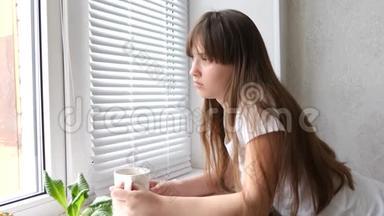 十几岁的女孩在靠近窗户的窗台上<strong>喝茶</strong>。 美丽悲伤的女孩望着窗外<strong>喝茶</strong>或咖啡。