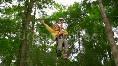 一个小男孩在森<strong>林探</strong>险公园的树梢上的高空滑索上穿着安全装备的慢速镜头。 室外