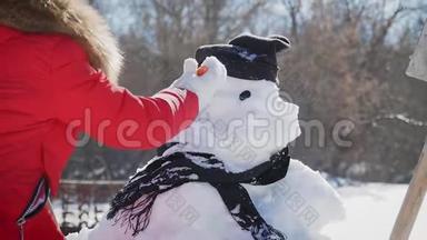 女孩在雪人上插入一个鼻子。 孩子玩雪人.. 冬天的快乐时光，雪上的孩子。