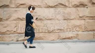 快乐的年轻女士戴着耳机听着音乐走在街上，双手握着移动的手