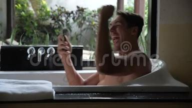 年轻英俊的男人躺在浴缸里，用智能手机，成功地交易，微笑。 男人在智能手机上获得胜利信息