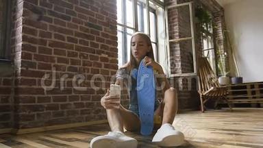 迷人的少女正坐在地板上，手里拿着滑板，在室内的智能手机上制作自拍照片