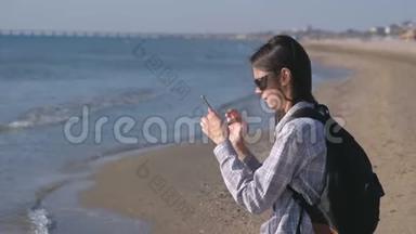 旅行者女孩和一个背包博主在沙滩上，并在手机上制作照片和视频。