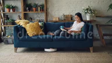 美丽的亚洲女孩正在看书，躺在客厅的沙发上，而机器人<strong>吸尘器</strong>则在干洗地板上