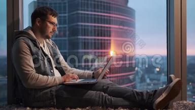一个年轻人坐在一台笔记本电脑后面，坐在一个地板上的窗户旁的画像