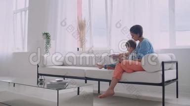 一位年轻的母亲带着一个孩子读一本书，坐在客厅里明亮的白色屋子里