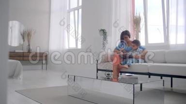 一位年轻的母亲带着一个孩子读一本书，坐在客厅里明亮的白色<strong>屋子</strong>里