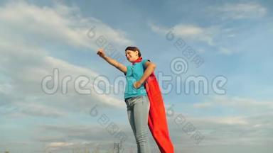 美丽的女孩超级英雄穿着红色斗篷站在田野上，斗篷随风飘扬。 慢动作。 特写镜头。 女孩的梦想