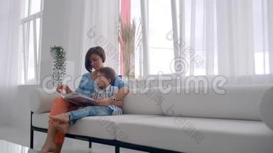 一位年轻的母亲带着一个孩子读一本书，坐在客厅里明亮的白色<strong>屋子</strong>里
