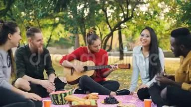 漂亮的女吉他手正在野餐中弹吉他，她的朋友们坐在野餐中唱歌，拍手