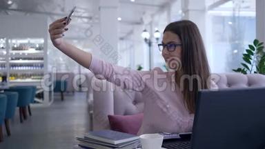 漂亮的餐馆老板女孩在笔记本电脑工作期间用手机拍自拍照片，并在笔记本上做笔记