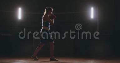 一个漂亮的女拳击手在黑暗的健身房训练，并在缓慢的运动中锻炼出拳头。 相机运动侧视图。 Stadicam