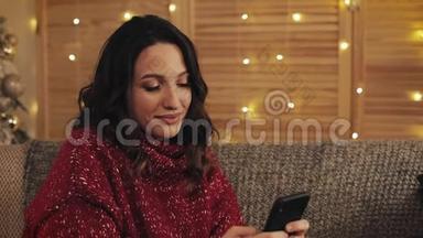 漂亮的女人使用智能手机坐在沙<strong>发</strong>上，<strong>发信息</strong>，在尼斯附近装饰的公寓里微笑。 圣诞树