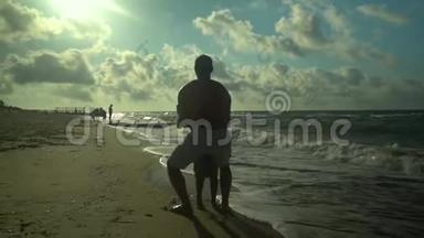 一位年轻的父亲和他的儿子沿着海滨散步。 阳光灿烂. 爸爸把他的儿子旋转，把他扔进水里