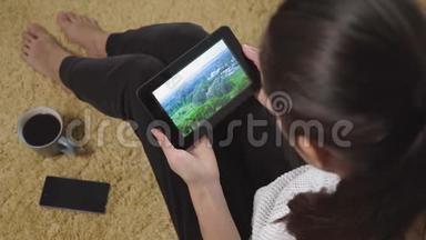 女人在地毯上坐在家里，用平板电脑在屏幕上观看大自然和度假视频