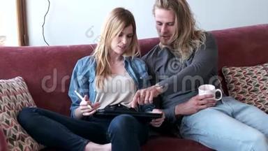 可爱的年轻夫妇在家坐在沙发上用数码平板电脑画画。