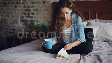 有着一头深色卷发的漂亮年轻女子正在看书翻页，端着杯子坐在床上欣赏文学作品