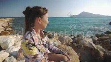 年轻迷人的女人微笑着坐在岩石上的夕阳山和海岸背景。 头发