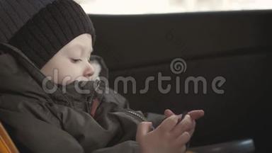 可爱的蹒跚学步的小男孩坐在汽车座椅上，从智能手机上观看视频。 小孩在车里玩智能手机
