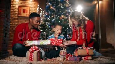 新年礼物中，一个幸福的家庭正坐在圣诞树`附近。 那个男孩正在用红色包装摇动礼物。