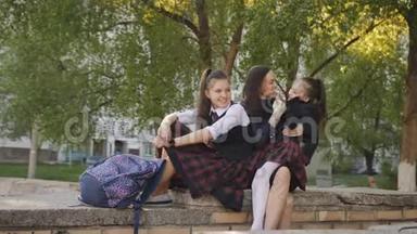 母亲带着两个穿校服的女儿，背包坐在学校附近的街道上，亲吻和交谈。