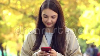 穿着外套或壕沟的年轻女子，用智能手机站在秋季公园里