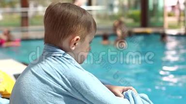 一个小男孩正坐在游泳池旁的一个阳光休息室里，用毛巾包着。