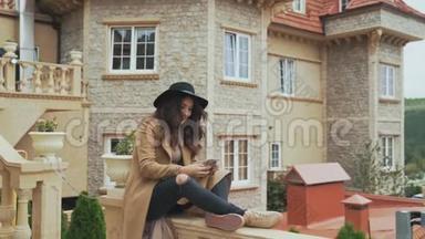 一位卷发的美女坐在一座古老的石头城堡的阳台栏杆上，给她的朋友写短信，用一个