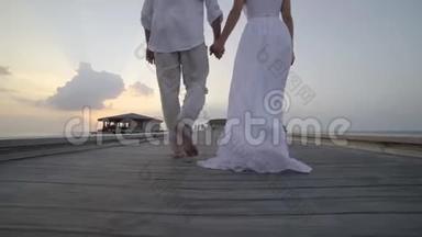 蜜月旅行在热带地区，新娘穿着白色礼服和新郎一起走在木制码头上，在豪华的平房里