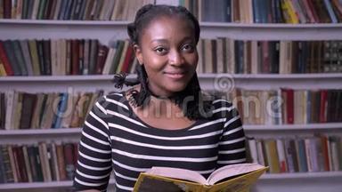 年轻美丽的非洲裔美国妇女在图书馆看书，微笑，快乐，书架背景