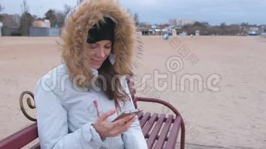 穿着白色<strong>羽绒服</strong>的女人坐在海滩上的长凳上，看着手机看着令人惊叹的视频。