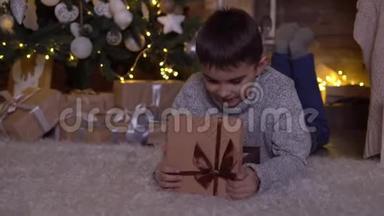 <strong>一个</strong>小男孩打开<strong>一个</strong>带礼物的盒子，高兴地躺在圣诞树附近的<strong>地板上</strong>。 4K