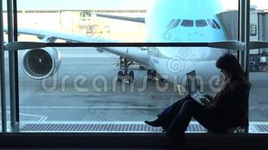 美丽的女人在机场看着笔记本电脑，坐在大窗户上，飞机停在那里