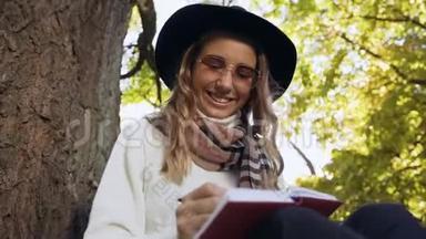可爱的金发女人戴着帽子坐在公园的树旁，在她的笔记簿中注意到了一些想法。
