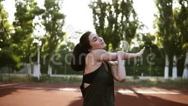 一个身穿运动服的活泼的黑发女孩伸展着她的双手肌肉，热身。 户外体育场，绿色