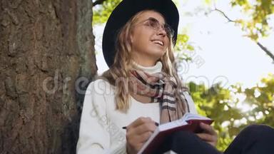 近距离拍摄年轻女子戴着时尚帽子，在笔记本上写一些东西，同时在城市公园放松。