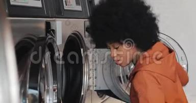 年轻的非洲裔美国妇女坐在洗衣机前，给洗衣机装上脏衣服。 自助服务
