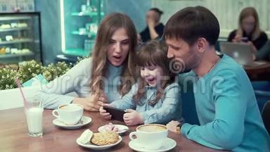 一家人在咖啡馆里，爸爸，妈妈，女儿在桌子上看着电话里的东西，微笑着。