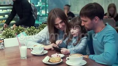一家人在咖啡馆里，爸爸，妈妈，女儿在桌子上看着电话里的东西，微笑着。