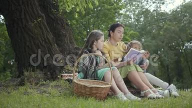 优雅的高级女人坐在公园树下的毯子上看书。 两个可爱的孙女坐着