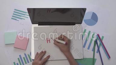 女人在笔记本电脑上的一张纸上写HACKING。 带有图表的办公桌面