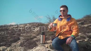一个穿着黄色夹克，蓝色牛仔裤和眼镜的男人坐在山上，欣赏风景，喝热水瓶里的茶。