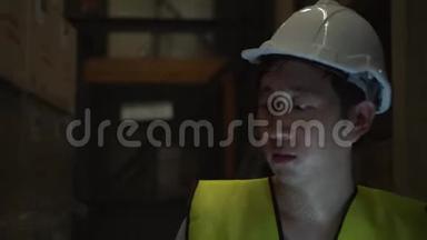 年轻的仓库工人戴着安全安全帽，带着数字平板电脑穿过库存室