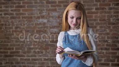 年轻感兴趣的金发女孩<strong>正在看书</strong>，看着摄像机，微笑着，带着砖头背景