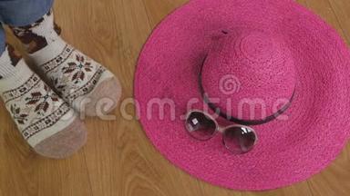 一个女人的腿坐在地板上，穿着温暖的羊毛袜子。 附近有一顶宽边的粉红色帽子和太阳镜