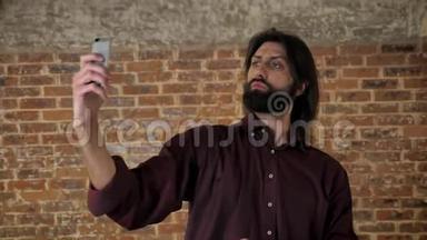 年轻迷人的黑发男子，蓄着胡须，正在智能手机上制作视频，在周围闲逛，笑，砖背景