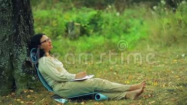 一个戴着眼镜的年轻微笑女子赤着脚<strong>坐在</strong>公园的一棵<strong>树下</strong>，在笔记本上画一支铅笔