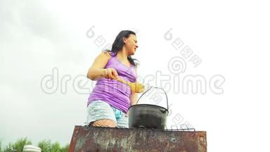 一个女人在火上用木勺在水壶里搅拌汤。 他远离烟雾。
