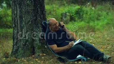 一个戴着眼镜的微笑男子<strong>坐在</strong>公园的一棵<strong>树下</strong>，用铅笔画一个记事本，然后用电话交谈